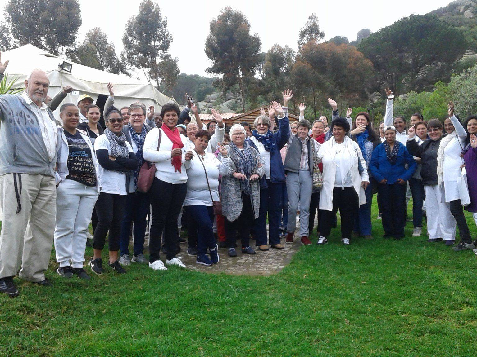 A large group of individuals standing together outdoors, smiling and waving at the camera.