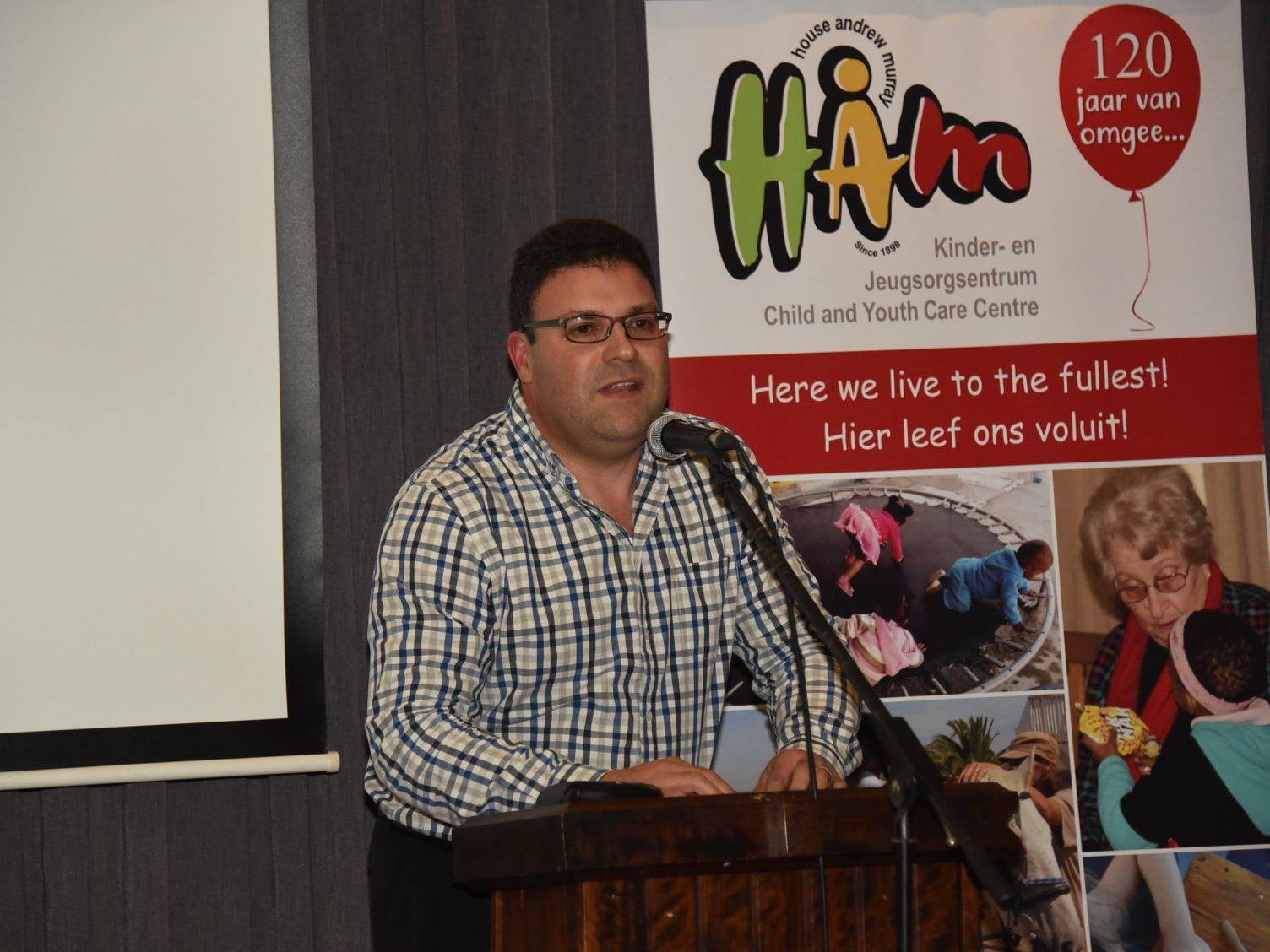 Man in a checkered shirt speaking at a podium during an event at a child and youth care centre.