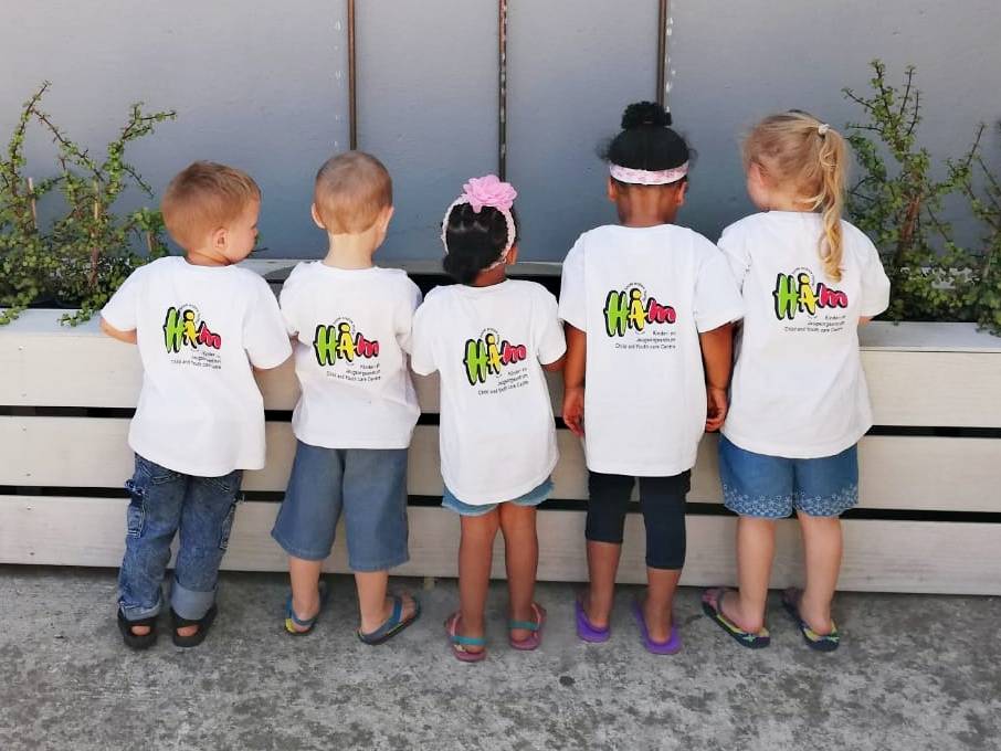 Five children standing in a row wearing white t-shirts with colorful logos on the back.